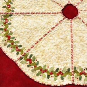 May include: Cream-colored quilted Christmas tree skirt with a red center and a decorative border. The border features green leaves, red berries, and red and white striped trim. The skirt has a scalloped edge.