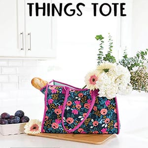 May include: A floral tote bag with pink handles and a dark blue background. The bag is sitting on a wooden surface with a loaf of bread and a bowl of plums in the background. The text "ALL THE THINGS TOTE" is displayed above the bag. The text "KNOT + THREAD design" is displayed below the bag.
