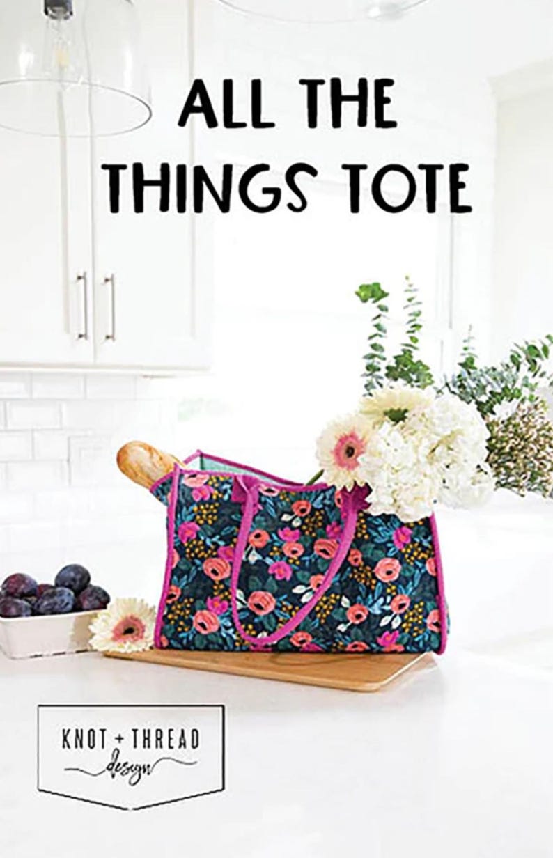 May include: A floral tote bag with pink handles and a dark blue background. The bag is sitting on a wooden surface with a loaf of bread and a bowl of plums in the background. The text "ALL THE THINGS TOTE" is displayed above the bag. The text "KNOT + THREAD design" is displayed below the bag.