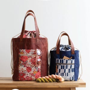 May include: Two drawstring tote bags with leather handles and floral and geometric pocket designs. The larger bag is brown with a floral pocket, and the smaller bag is navy blue with a geometric pocket. The text "FIREFLY TOTE" is at the top.