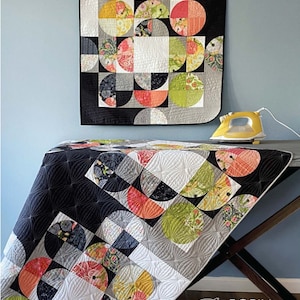 May include: A quilt with a geometric design featuring half-circle patterns in various colors, including black, white, gray, orange, and green. The quilt is displayed on a table with an iron. The text "Partial Eclipse" is at the top.