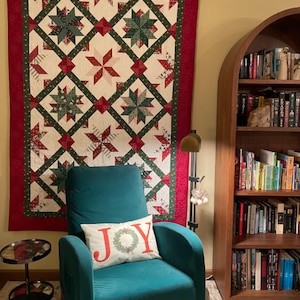 May include: A Christmas-themed quilt with red and green star patterns hangs above a teal armchair. A pillow with the word "JOY" is on the chair. A dark wooden bookshelf filled with books is to the right. A small side table is to the left.