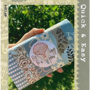 May include: A travel companion with a patchwork design in shades of blue, gray, and cream, featuring floral and botanical patterns. A circular patch with a hot air balloon and the text "Fly to a happy place" is visible. The text "Fly to Anywhere TRAVEL COMPANION" is also visible.