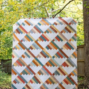 May include: A large, handmade quilt with a geometric pattern. The quilt features a repeating design of angled stripes in shades of orange, teal, brown, and cream. The quilt is displayed outdoors, with a natural background of trees and foliage.