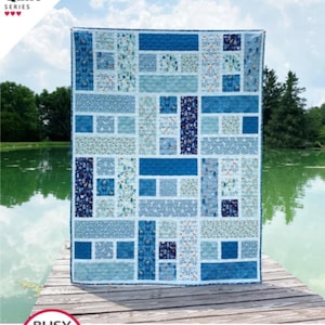 May include: A blue and white quilt with a rectangular block design. The quilt is displayed outdoors on a wooden dock with a lake in the background. The text "Blessings" is at the top.