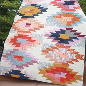 May include: A colorful quilt with a geometric design. The quilt features a repeating pattern of angular shapes in shades of orange, pink, blue, and yellow on a cream background. The quilt measures 80.5 inches by 80.5 inches. The word "Weftovers" is printed on the quilt.