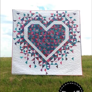 May include: A white quilt with a large pink and blue heart design. The heart is surrounded by smaller triangles in shades of pink, blue, and purple. The quilt is displayed outdoors on a grassy field. The text "Exploding Heart" is at the top of the image and "Slice of Pi Quilts" is at the bottom.