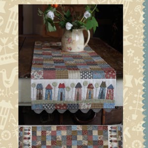 May include: A patchwork table runner with a "Tiny Town" design. The runner features a repeating pattern of small houses and a checkered pattern in shades of blue, red, and brown. The top of the image shows a vase with white flowers.