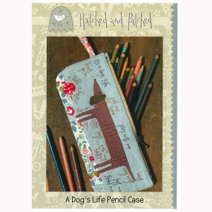 May include: A Dog's Life pencil case with a brown dog illustration and text. The pencil case is light blue with a floral pattern on the side. The case is filled with various colored pencils.