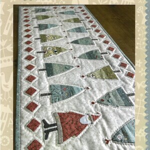 May include: A quilted Christmas tree table runner with a repeating pattern of Christmas trees and Santa Claus figures. The trees are various shades of green and blue, and the Santa figures are red. The table runner is bordered with red diamond shapes.