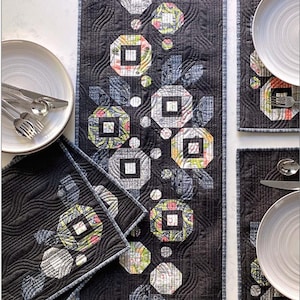 May include: A set of four black and white quilted placemats and a table runner with a geometric pattern of squares and circles. The pattern features a floral design in shades of pink, green, and yellow. The text "POSIES TABLE RUNNER AND 4 PLACEMATS" is printed in white on a red background at the top of the image. The text "ROBIN PICKENS QUILT PATTERNS" is printed in white on a red background at the bottom of the image.