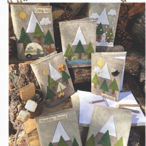 May include: A set of six felt notebook covers with a woodland theme. Each cover features a different design with mountains, trees, and other nature-inspired elements. The covers are labeled with titles such as "Camp Log", "Mountain Journal", and "Field Guide".
