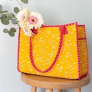 May include: A yellow quilted tote bag with pink and red checkered trim and daisy print. The bag has two handles and is sitting on a wooden stool.