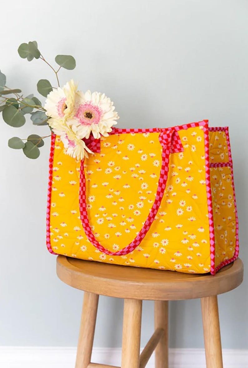 May include: A yellow quilted tote bag with pink and red checkered trim and daisy print. The bag has two handles and is sitting on a wooden stool.