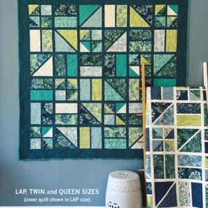 May include: A teal and green quilt with geometric patterns, including triangles and rectangles, hangs on a wall. The quilt is displayed with a matching smaller quilt. The text "BACKSLASH" is at the top, and "LAP, TWIN and QUEEN SIZES" is at the bottom.