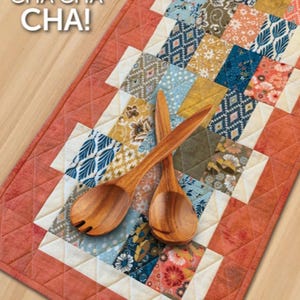 May include: A colorful patchwork table runner with a red border and a white inner border. The runner is made of various fabrics with floral and geometric patterns. Two wooden spoons are placed on top of the runner.