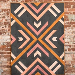 May include: A geometric quilt with a dark gray background and a pattern of orange, pink, and white chevron shapes. The quilt is rectangular and has a brown border. The quilt is hanging on a brick wall.