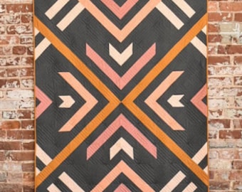 May include: A geometric quilt with a dark gray background and a pattern of orange, pink, and white chevron shapes. The quilt is rectangular and has a brown border. The quilt is hanging on a brick wall.