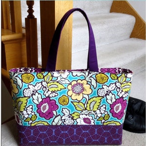 May include: The Essential Tote bag with a floral pattern in turquoise, yellow, and purple. The bag has a purple handle and a geometric patterned base. The text on the bag reads "The Essential Tote" and "A Beginner Bag with a Sophisticated Look". The bag is approximately 12-3/4 inches high, 19-1/4 inches wide, and 4 inches deep.