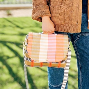 May include: A colorful, striped handbag with a flap closure. The bag features a mix of orange, pink, white, and yellow stripes. It has a handle and a detachable striped shoulder strap. The bag is held by a person wearing a brown cardigan and blue jeans.