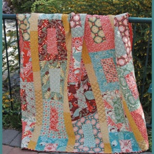 May include: A colorful quilt pattern called "Penny Lane" by Abbey Lane Quilts. The quilt is made with a variety of fabrics in shades of red, orange, yellow, green, blue, and brown. The quilt is hanging on a black metal railing.