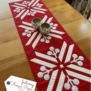 May include: Red and white table runner with a snowflake design and white berries. The table runner is 13 1/2 inches by 40 inches and features freezer paper piecing. "Snowberry Runners" and "featuring Freezer Paper Piecing!" are written on the image.