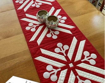 Può includere: Un runner da tavolo rosso e bianco con un motivo a fiocco di neve e bacche bianche. Il runner da tavolo misura 34 cm x 101,6 cm e presenta la tecnica del "freezer paper piecing". "Snowberry Runners" e "featuring Freezer Paper Piecing!" sono scritti sull'immagine.