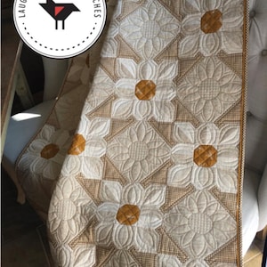 May include: A beige and brown quilted wall hanging or lap quilt with a daisy pattern. The quilt is 45 inches by 45 inches or 45 inches by 63 inches. The quilt is made with a beige fabric with a brown gingham pattern and has a brown border. The quilt is quilted with a daisy pattern in a white fabric. The quilt is on a white chair with a brown and white patterned fabric. The quilt has the text "Laugh Yourself Into Stitches" on a black circle with a red bird inside.