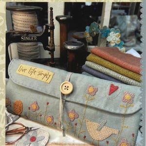 May include: A light blue sewing case with embroidered flowers and a bird design. The case has a button closure and the text "Live life simply". The words "Cherish Each Day Sewing Case" are at the bottom. A Singer sewing machine is in the background.