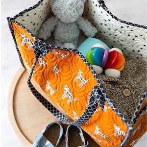 May include: A large orange and black quilted tote bag with a zipper closure. The bag is filled with a gray stuffed animal, a blue and white blanket, and a colorful toy. Two pairs of blue denim shoes are sitting on a wooden surface.
