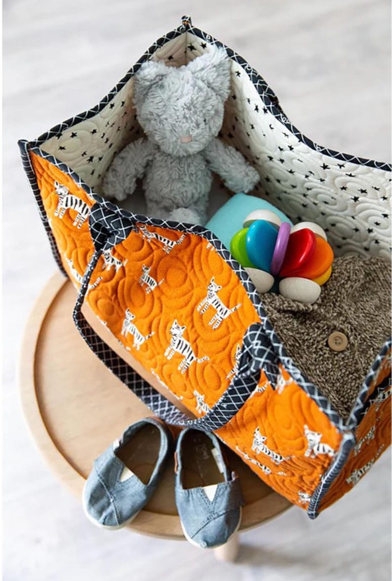 May include: A large orange and black quilted tote bag with a zipper closure. The bag is filled with a gray stuffed animal, a blue and white blanket, and a colorful toy. Two pairs of blue denim shoes are sitting on a wooden surface.
