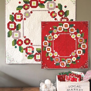 May include: Two quilts with a floral wreath design, one on a white background and the other on red. The quilts are labeled "Little Posy Wreath" and are available in 32.5" or 48.5" square sizes. A wooden box labeled "Local Market" and a glass bowl are in the foreground.
