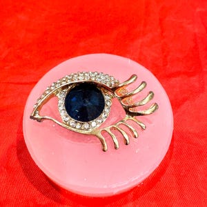 May include: A gold eye-shaped brooch with a blue rhinestone center and clear rhinestones around the eye. The brooch is on a pink background.