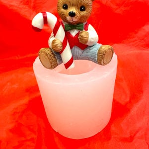 May include: A white ceramic candle holder with a brown teddy bear figurine holding a red and white candy cane. The bear is wearing a red vest and blue pants with a green bow tie.