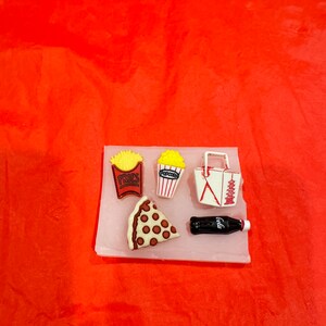 Puede incluir: Un molde de silicona blanco con cinco cavidades con forma de comida. Las cavidades tienen forma de caja de patatas fritas, caja de palomitas de maíz, porción de pizza, contenedor para llevar y botella de cola. El molde está sobre una superficie roja.