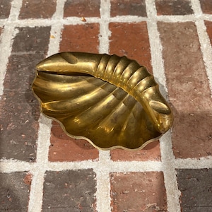May include: A gold-colored, shell-shaped dish with a textured surface. The dish has a scalloped edge and is resting on a brick surface. The dish is likely used for decorative purposes or to hold small items.