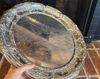 May include: A silver serving tray with an ornate, embossed design around the edge. The tray is round and has a smooth, reflective surface.