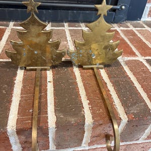 May include: A pair of brass stocking holders, each featuring a flat, stylized Christmas tree silhouette topped with a star. The metal surfaces show signs of oxidation and wear, and each tree is attached to a long, curved hook for mantel display.