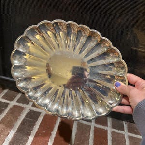 May include: A silver-colored, scalloped-edge decorative plate. The plate has a sunburst design radiating from the center. The plate is held by a person with a purple painted fingernail.