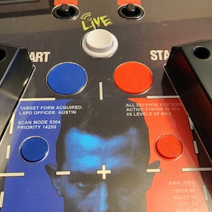 May include: A black and blue arcade game control panel with red and blue buttons. The panel has text that reads "TARGET FORM ACQUIRED: LAPD OFFICER: AUSTIN SCAN MODE 0304 PRIORITY 14250" and "ALL DEFENSE SYSTEMS ACTIVE STATUS 56 85A I/O LEVELS AT MAX".