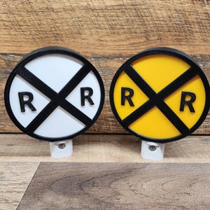May include: Two round railroad crossing signs with black 'RR' letters on a white background and a yellow background. The signs are mounted on white bases and are illuminated by a small light bulb.