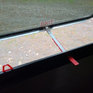 May include: A close-up of a window with a screen and a metal latch. The window is open and the latch is in the open position. The window has a red label that says "EXIT".