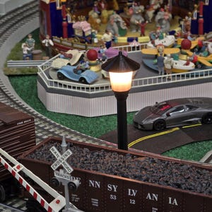 May include: A model train set with a brown boxcar, a black and silver sports car, and a streetlight with a white light. The train set is on a track with a railroad crossing sign. The sports car is on a road with yellow lines. The streetlight is on a black pole.