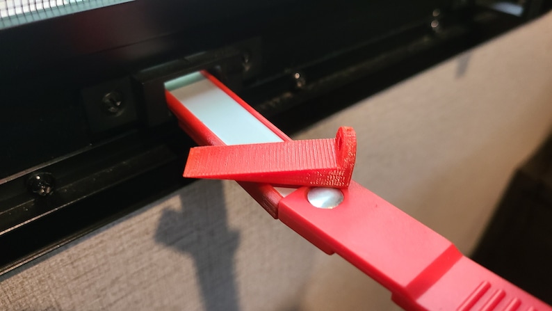 May include: A red plastic window latch with a white strip and a small metal piece. The latch is attached to a black window frame.
