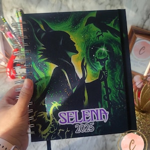 May include: A black spiral bound notebook with a green and black image of Maleficent from Disney's Sleeping Beauty. The image includes Maleficent's silhouette, her raven, and her staff. The notebook has the text "SELENA 2025" printed on the front.