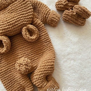 May include: A brown, knitted baby outfit set, including a one-piece suit with attached ears and a pom-pom, and two matching booties. The set is made of a soft, textured yarn and is displayed on a white, fluffy surface.