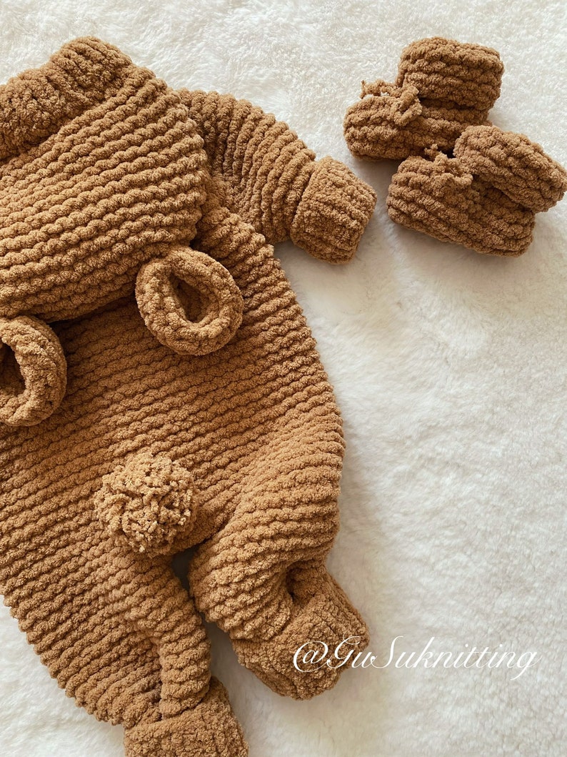 May include: A brown, knitted baby outfit set, including a one-piece suit with attached ears and a pom-pom, and two matching booties. The set is made of a soft, textured yarn and is displayed on a white, fluffy surface.
