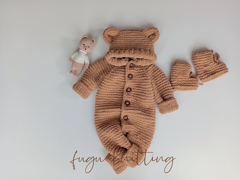 May include: A tan-colored, crocheted baby outfit set, including a hooded jumpsuit with bear ears, matching booties, and a small teddy bear toy. The jumpsuit has wooden buttons down the front. The text "fugu knitting" is at the bottom.
