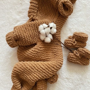 May include: A brown, knitted baby outfit with a hood and matching booties. The outfit is made of a soft, textured yarn and features a bear-like design. A sprig of cotton adds a delicate touch to the presentation. The outfit is designed for a baby.