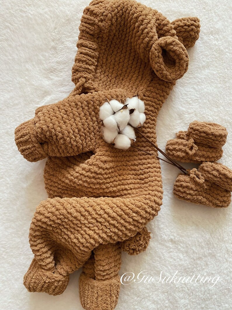 May include: A brown, knitted baby outfit with a hood and matching booties. The outfit is made of a soft, textured yarn and features a bear-like design. A sprig of cotton adds a delicate touch to the presentation. The outfit is designed for a baby.